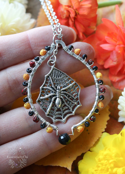 Silver Beaded Hoop Necklace with a spider on a web charm and a faceted black tourmaline focal bead. The necklace is beaded with a gradient of orange, blood red, and matte black glass beads. A stylish and mystical accessory, perfect for adding a touch of gothic and witchy flair to your outfit.
