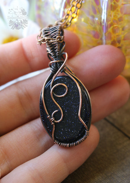 Handcrafted Blue Goldstone Copper Wire Wrapped Pendant - A shimmering gemstone pendant with a mesmerizing night-sky appearance, expertly wrapped in copper wire. This unique piece combines the cosmic allure of Blue Goldstone with the rustic charm of artisan craftsmanship, creating a striking and versatile accessory for crystal enthusiasts. Showing the back of the pendant and that it's pretty swirls on the back are just as pretty as the front.