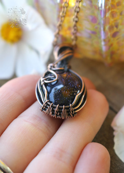 Handcrafted Blue Goldstone Copper Wire Wrapped Pendant - A shimmering gemstone pendant with a mesmerizing night-sky appearance, expertly wrapped in copper wire. This unique piece combines the cosmic allure of Blue Goldstone with the rustic charm of artisan craftsmanship, creating a striking and versatile accessory for crystal enthusiasts. Showing the base of the pendant and the intricate weaving pattern.