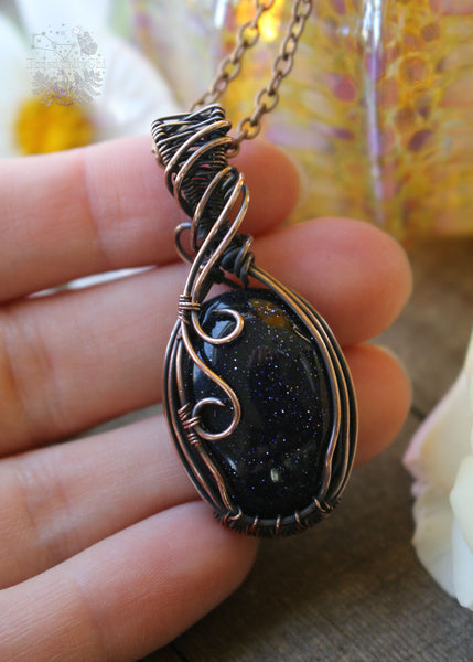Handcrafted Blue Goldstone Copper Wire Wrapped Pendant - A shimmering gemstone pendant with a mesmerizing night-sky appearance, expertly wrapped in copper wire. This unique piece combines the cosmic allure of Blue Goldstone with the rustic charm of artisan craftsmanship, creating a striking and versatile accessory for crystal enthusiasts. Shown in hand for size reference.