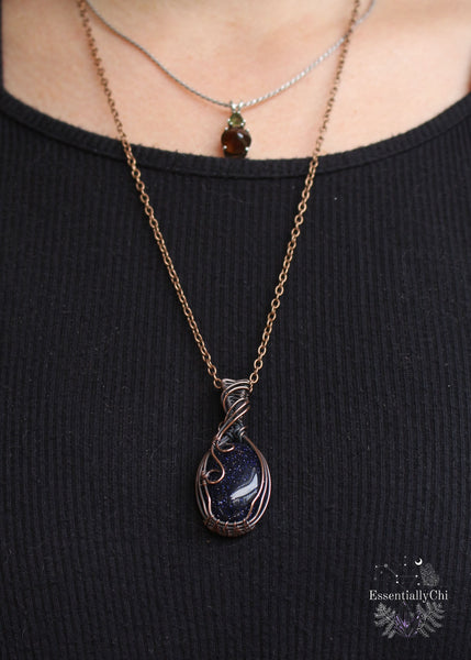Handcrafted Blue Goldstone Copper Wire Wrapped Pendant - A shimmering gemstone pendant with a mesmerizing night-sky appearance, expertly wrapped in copper wire. This unique piece combines the cosmic allure of Blue Goldstone with the rustic charm of artisan craftsmanship, creating a striking and versatile accessory for crystal enthusiasts. Shown worn to see how it hangs along the neckline. Hangs at above the heart space.