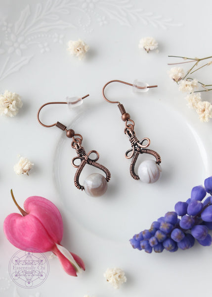 Handcrafted Lily earrings featuring Botswana agate stones wrapped in copper wire. The shape of the earrings is a pear shape with two loops above the stone that looks like a bow. The color of the Botswana agate bead banding is grey and white. Copper wire is antiqued to bring out the details.