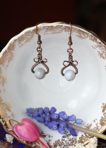 Handcrafted Lily earrings featuring Botswana agate stones wrapped in copper wire. The shape of the earrings is a pear shape with two loops above the stone that looks like a bow. The color of the Botswana agate bead banding is grey and white. Copper wire is antiqued to bring out the details.
