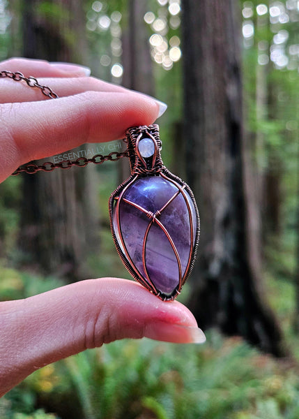 Chevron Amethyst and Rainbow Moonstone copper wire-wrapped jewelry piece with filigree details. Chevron Amethyst criss-cross design on the front, and a blue flash Rainbow Moonstone in the bail. Back features intricate filigree design. Held up showing the redwood forest behind it.