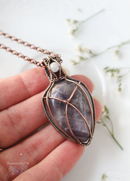 Athena pendant, a Chevron Amethyst and Rainbow Moonstone copper wire-wrapped jewelry piece with filigree details. Chevron Amethyst criss-cross design on the front, symbolizing Athena's strategic strength, and a blue flash Rainbow Moonstone in the bail. Back features intricate filigree for security and elegance, reminiscent of Athena's wisdom and warrior spirit.