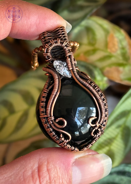 Rainbow Obsidian sphere wrapped in copper wire and an accent diamond quartz in the bail, elegantly wrapped. The Sphere has a green and slightly purple sheen. Shown held in hand for size reference.