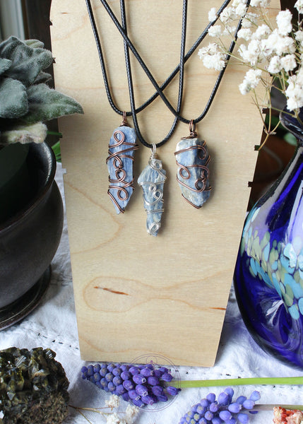 Three wire wrapped raw blue kyanite necklaces on a black waxed cotton cord shown hanging. First one is copper wire, second is silver plated, third is copper wire and a little more chunky. Raw Blue Kyanite Wire Wrapped Necklace for energy protection, featuring a simple, everyday wear design that enhances communication, alignment, tranquility, restful sleep, and dream recall.