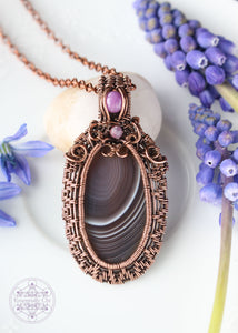 Handcrafted Primrose pendant featuring a grey banded Botswana Agate stone framed with woven copper wire frame and a filigree design around the base of the bail. Includes accent stones of phosphosiderite and watermelon tourmaline.