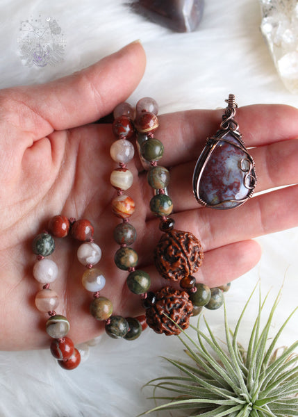 Longing for the serenity of a forest walk? Let the Amanita Forest Mala carry you away. Hand-knotted with Flower Agate, Brecciated Red Jasper, Rainforest Jasper, Smoky Quartz, and 4 Rudraksha seeds, this 72-bead mala necklace is designed to help nature lovers, energy healers, and nature witches ground themselves and open their hearts to new opportunities. Crafted with a copper wire-wrapped Red Moss Agate pendant, this unique piece is the perfect companion for your meditation practice. Beaded Mala