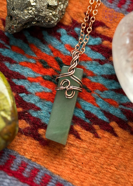 Green Aventurine Copper wire wrapped necklace with a a simple design in the front that looks like hands clasping together and has a braided bail. Hangs on a black cotton wax cord. Shown on a navajo wool mat with crystals around it for display.
