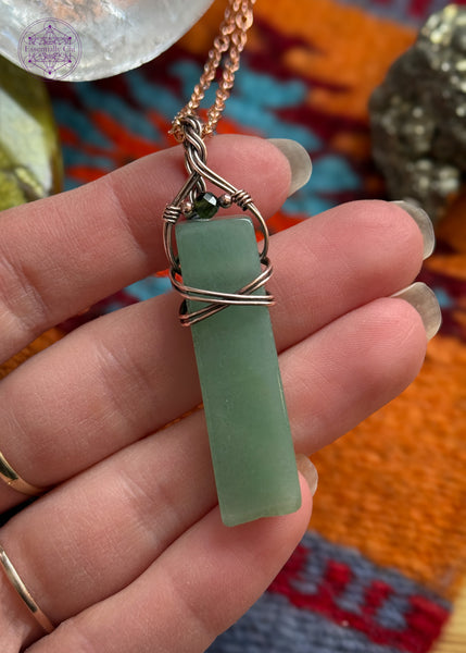 Green adventurine rectangular style pendant with a simple wrap of a criss cross over the face of the stone and a braided bail. an added green tourmaline stone is at the base of the bail. hung on a 22" black nylon cord. Held in hand to show size ratio.