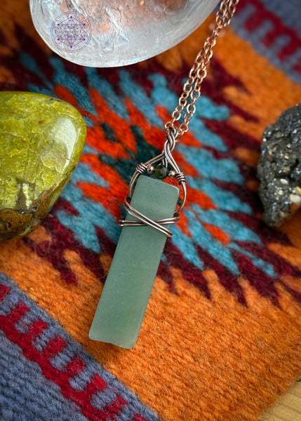 Green adventurine rectangular style pendant with a simple wrap of a criss cross over the face of the stone and a braided bail. an added green tourmaline stone is at the base of the bail. hung on a 22" black nylon cord.