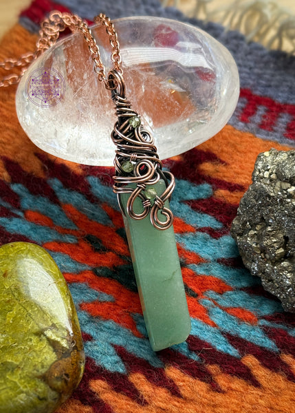 Green Aventurine rectangular crystal wrapped in copper wire around the bail with a swirly filagree design and peridot accent crystals placed within the design. Placed near crystals to display.