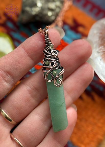 Green Aventurine rectangular crystal wrapped in copper wire around the bail with a swirly filagree design and peridot accent crystals placed within the design. Held in hand to show size.