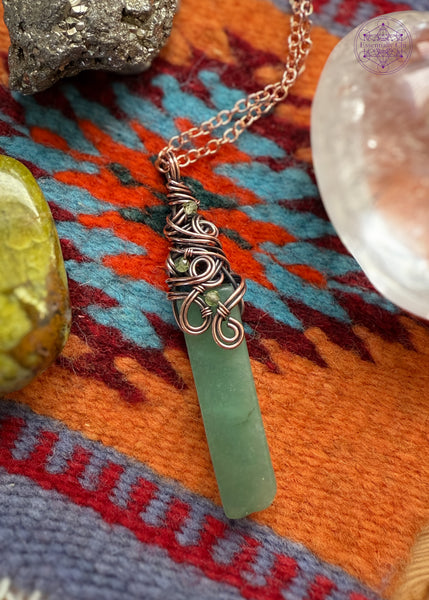 Green Aventurine rectangular crystal wrapped in copper wire around the bail with a swirly filagree design and peridot accent crystals placed within the design. Placed on a Navajo wool mat and crystals around it for display.