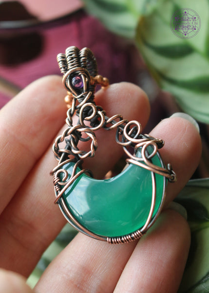 Green onyx moon crystal wrapped in copper wire to make it look like a bubbling cauldron. Bubble loops in the copper wrap around a small amethyst crystal at the base of the bail. Held in hand to show size. 1.9" long.