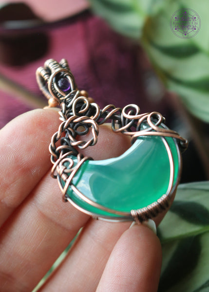 Green onyx moon crystal wrapped in copper wire to make it look like a bubbling cauldron. Bubble loops in the copper wrap around a small amethyst crystal at the base of the bail. Held in hand sideways to show detail.