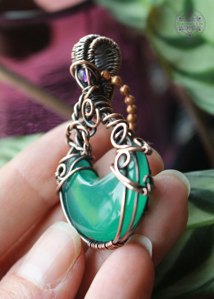 Green onyx moon crystal wrapped in copper wire to make it look like a bubbling cauldron. Bubble loops in the copper wrap around a small amethyst crystal at the base of the bail. Held in hand on the left side to show detail.