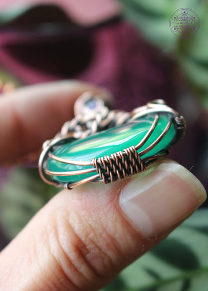 Green onyx moon crystal wrapped in copper wire to make it look like a bubbling cauldron. Bubble loops in the copper wrap around a small amethyst crystal at the base of the bail. Held in hand to show bottom wrap detail.