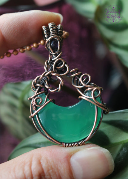 Green onyx moon crystal wrapped in copper wire to make it look like a bubbling cauldron. Bubble loops in the copper wrap around a small amethyst crystal at the base of the bail. Held in hand to show size. 1.9" long.