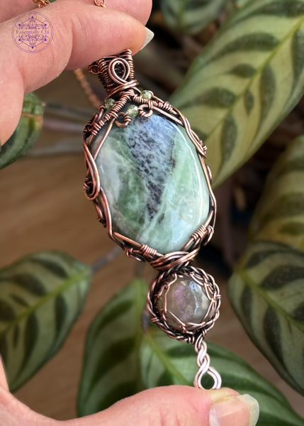 A copper wire wrapped crystal pendant with crystals of an oval Green Kammererite as the main stone, below it is a smaller round shaped purple labradorite, amd a braided twist design at the base. The kammererite has a filagree design with faceted peridot beads woven into the design. Held in hand to show size with a prayer plant behind it to bring out the greens in the crystal.