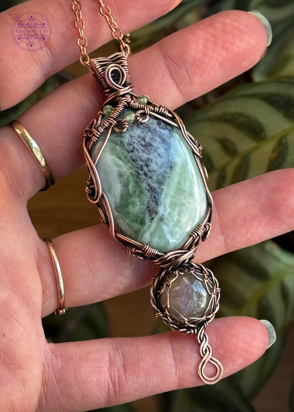 A copper wire wrapped crystal pendant with crystals of an oval Green Kammererite as the main stone, below it is a smaller round shaped purple labradorite, amd a braided twist design at the base. The kammererite has a filagree design with faceted peridot beads woven into the design. Held in hand to show size with a prayer plant behind it to bring out the greens in the crystal.