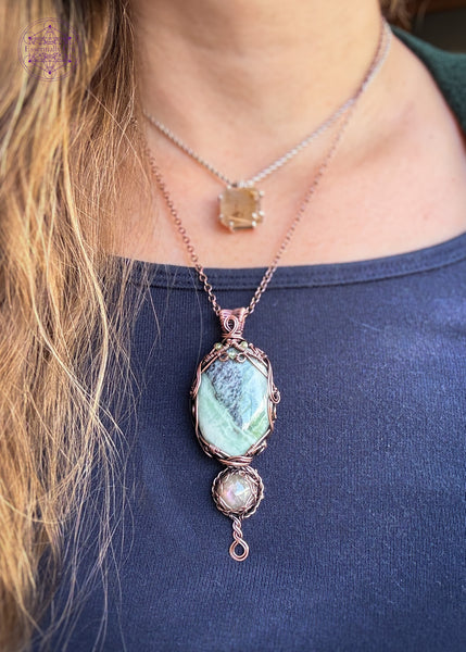 A copper wire wrapped crystal pendant with crystals of an oval Green Kammererite as the main stone, below it is a smaller round shaped purple labradorite, amd a braided twist design at the base. The kammererite has a filagree design with faceted peridot beads woven into the design. Shown worn around the neck to display size ratio.