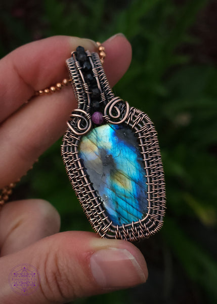 Copper wire wrapped labradorite coffin pendant with a red tourmaline accent stone at the top of the coffin to symbolize blood color, and then black spinel beads along the center of the bail. nice thick wrap style to create the ultimate statement piece. Labradorite has mainly blue flash with a few dark spots and yellow to orange colors. Held in hand for size reference.