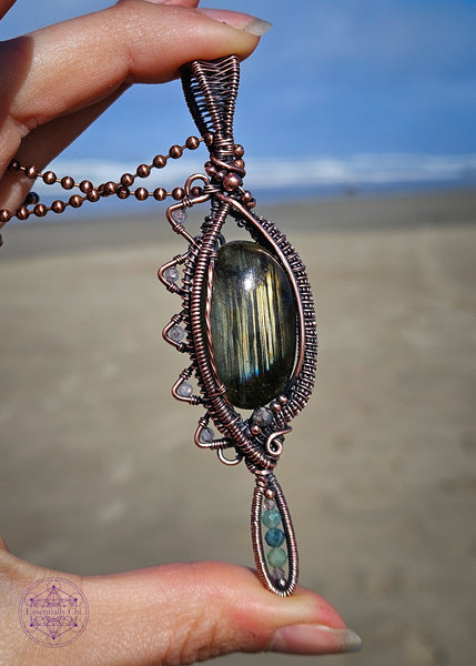 Copper wire wrapped necklace with an oval labradorite that has lines of yellow, green and blue flashes like the Norther Lights. On the left side there is a zig zag pattern with faceted rainbow moonstone bead with blue flash. The zig zag makes the pendant look like an eye when held sideways. At the base of the pendant is a long oval loop that encases small faceted smoky quartz and green tourmaline beads. Theres a copper coiling up the right side and copper accent beads throughout, truly a statement piece!