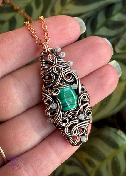 Malachite wrapped in a diamond shaped copper wire frame with a filagree design and swirls, with added accents of 2mm rainbow moonstone faceted beads. Held in hand to show size reference.