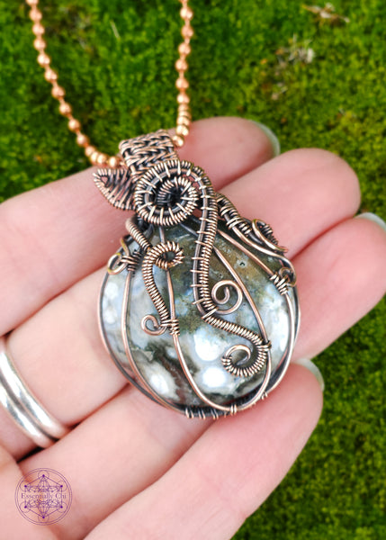 Copper wire wrapped pumpkin pendant with a green ocean jasper crystal. The white dots on the ocean jasper remind me of the warts on the warted pumpkins for this design. The bail is short to resemble a stem, and a wire wrapped leaf out the base of the stem. Vine curls around the ridges of the pumpkin. Shown in hand for size.