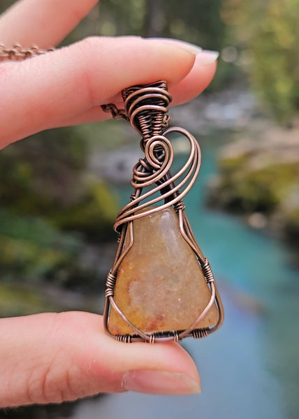 Orange Aventurine copper wire wrapped necklace shown in hand with a background of aqua turquoise water from Mount Rainier glaciers.