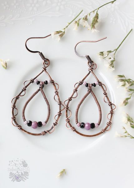 A pair of elegant tear-drop-shaped earrings with intricate filigree designs along the edges. These eye-catching earrings feature an additional tear-drop dangle inside, adorned with delicate pink and black seed beads. At the center of each earring, a faceted Phosphosiderite bead adds a touch of sophistication and metaphysical charm. The combination of filigree detailing, seed beads, and the unique gemstone creates a stylish and distinctive accessory, perfect for adding a touch of elegance to any outfit.