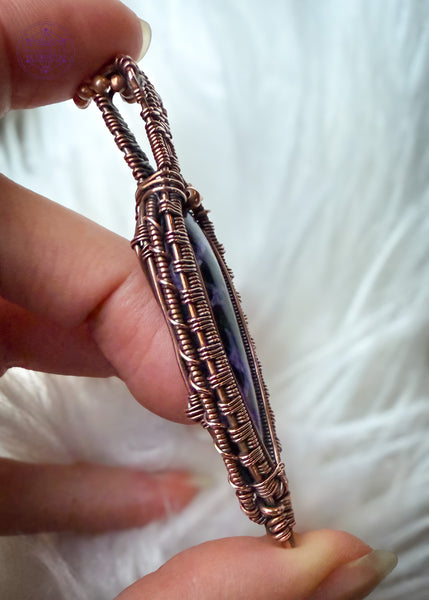 Copper wire wrapped Charoite pendant held in hand over a faux white fur. Showing the side of the pendant to show the detail of the coiling design on the sides. The bright light shows the detail in the stone that has some lavender chantoyancy and dark black inclusions. The bail has two faceted smoky quartz beads tucked into two triangles making this look like a galatic design.