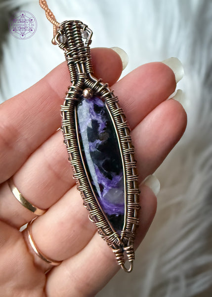 Copper wire wrapped Charoite pendant held in hand over a faux white fur. The bright light shows the detail in the stone that has some lavender chantoyancy and dark black inclusions. The bail has two faceted smoky quartz beads tucked into two triangles making this look like a galatic design.