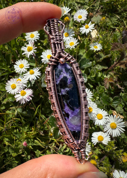 Copper wire wrapped Charoite pendant held in hand over green grass and tiny daisiys in the background. The bright light shows the detail in the stone that has some lavender chantoyancy and dark black inclusions. The bail has two faceted smoky quartz beads tucked into two triangles making this look like a galatic design.