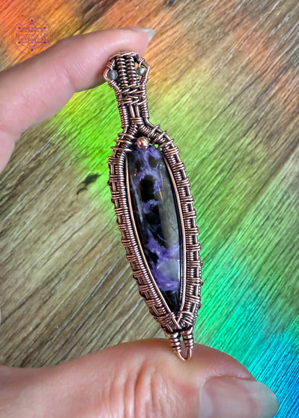 Copper wire wrapped Charoite pendant held in hand over a wood flooring and a rainbow steak from my window cling. The simplicity of the floor shows the detail in the wrap. The charoite stone has some lavender chantoyancy and dark black inclusions. The bail has two faceted smoky quartz beads tucked into two triangles making this look like a galatic design.