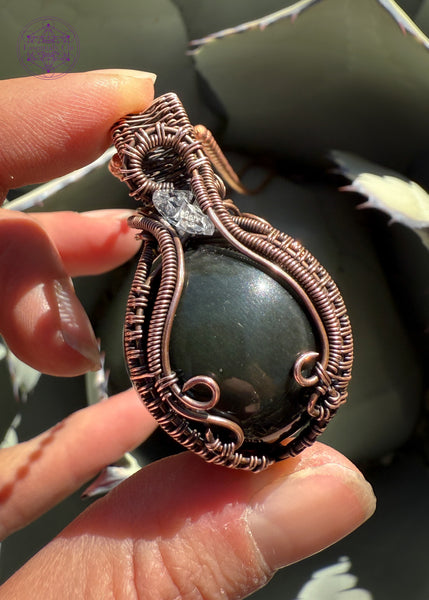 Rainbow Obsidian sphere wrapped in copper wire and an accent diamond quartz in the bail, elegantly wrapped. The Sphere has a green and slightly purple sheen. Held in hand to show size. Shown in bright light to show the green sheen in the shere.