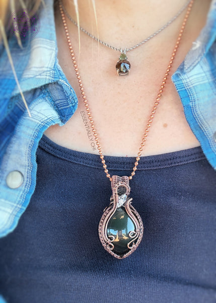 Rainbow Obsidian sphere wrapped in copper wire and an accent diamond quartz in the bail, elegantly wrapped. The Sphere has a green and slightly purple sheen.Shown being worn to see how it hangs on the neck.