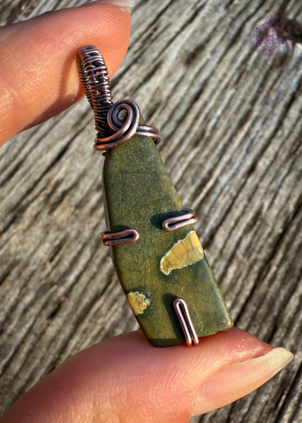 This rainforest rhyolite pendant is wrapped in copper wire with a simple prong design, one prong at the base, and one on each side. The bail is a simple narrow bail with a delicate swirl at the base to hold the pendant together. Simple elegant olive green stone with map like creamy yellow spots within the stone. Shown in hand for size reference and with a aged wooden background.