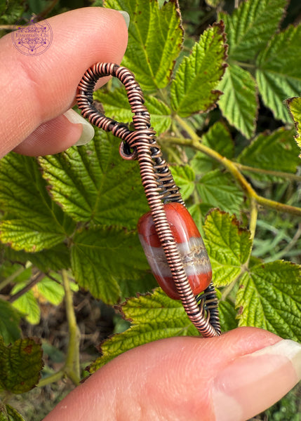 Copper wire wrapped red jasper bead with a river of quartz running in the middle of the jasper. Gorgeous bead centered in a minimalist design that kinda looks like a snow shoe. The bail is a simple criss cross woven design and the body of the pendant has a criss cross design centering the bead. Showing the side for reference, the back of the bead touches the skin and is not covered in wire.