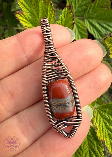 Copper wire wrapped red jasper bead with a river of quartz running in the middle of the jasper. Gorgeous bead centered in a minimalist design that kinda looks like a snow shoe. The bail is a simple criss cross woven design and the body of the pendant has a criss cross design centering the bead. held in hand for size reference.
