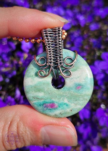 back of the Copper wire wrapped pendant with a donut shaped ruby fuchsite crystal that freely spins if you want a fidget spinner. The ruby in the fuchsite glows under a UV light. Shown in hand for size reference in front of purple flowers for a background.