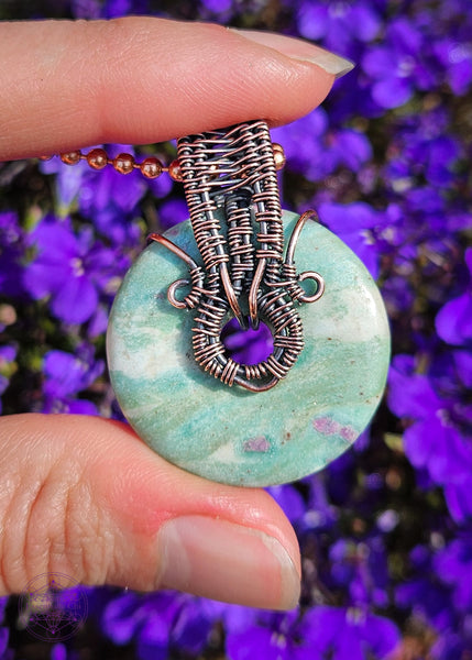 front of the Copper wire wrapped pendant with a donut shaped ruby fuchsite crystal that freely spins if you want a fidget spinner. The ruby in the fuchsite glows under a UV light. Shown in hand for size reference in front of purple flowers for a background.