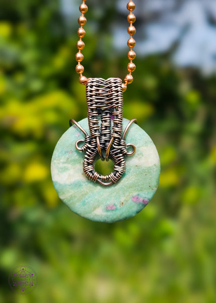 Copper wire wrapped pendant with a donut shaped ruby fuchsite crystal that freely spins if you want a fidget spinner. The ruby in the fuchsite glows under a UV light. hanging from a copper chain and shown in front of a green foliage background.