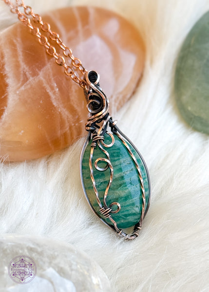 copper wire wrapped amazonite with a filagree design that swirls down the front right side of the oval crystal. This is a Russian Amazonite, so a darker aqua green than the paler Amazonite. This pendant is sitting on a faux white fur and crystals in the background for aesthetics.