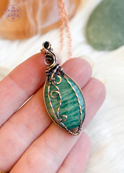 copper wire wrapped amazonite with a filagree design that swirls down the front right side of the oval crystal. This is a Russian Amazonite, so a darker aqua green than the paler Amazonite. Held in hand to show size reference.