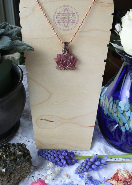 Tanzberry Lotus Copper Wire Wrapped Necklace featuring a carved lotus tanzberry that is very sparkly (red aventurine) crystal, wrapped in a simple copper wire that flows with the petals of the lotus. Pendant is 1.5" long on an 18" copper chain. Enhances determination, perseverance, and spiritual awakening. Shown on a wooden display to see how it hangs.