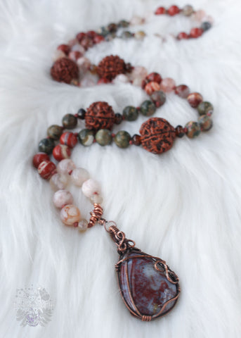Longing for the serenity of a forest walk? Let the Amanita Forest Mala carry you away. Hand-knotted with Flower Agate, Brecciated Red Jasper, Rainforest Jasper, Smoky Quartz, and 4 Rudraksha seeds, this 72-bead mala necklace is designed to help nature lovers, energy healers, and nature witches ground themselves and open their hearts to new opportunities. Crafted with a copper wire-wrapped Red Moss Agate pendant, this unique piece is the perfect companion for your meditation practice. Beaded Mala