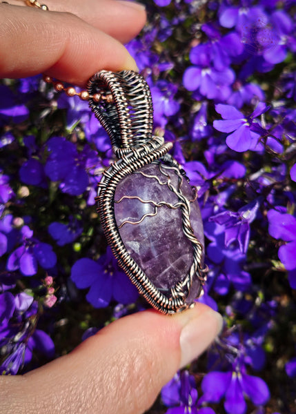 showing the left side of this copper wire wrapped amethyst tree of life pendant held in hand with purple lobelia flowers in the background. The Amethyst is a dark purple color and the roots have a swirl pattern near the bottom to symbolize the roots growing on rocks. There's a woven pattern along the sides of the pendant and a sturdy bail. 